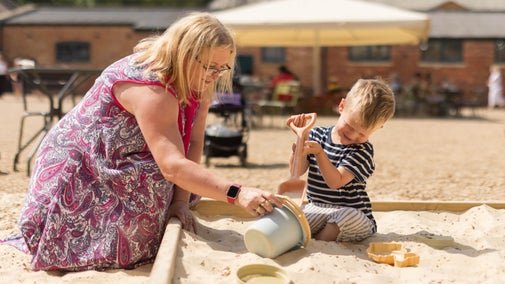 A woman and young child play together in a sand pit at Belton with a spade and bucket and plastic objects.